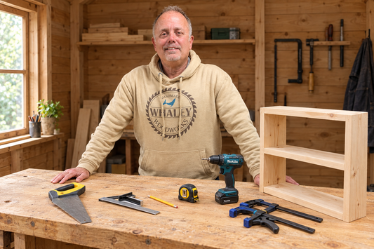 Jason wearing a Whaley Woodworks hoodie behind a workbench with basic woodworking tools and a small unfinished wooden shelf in a shed workshop.⠋