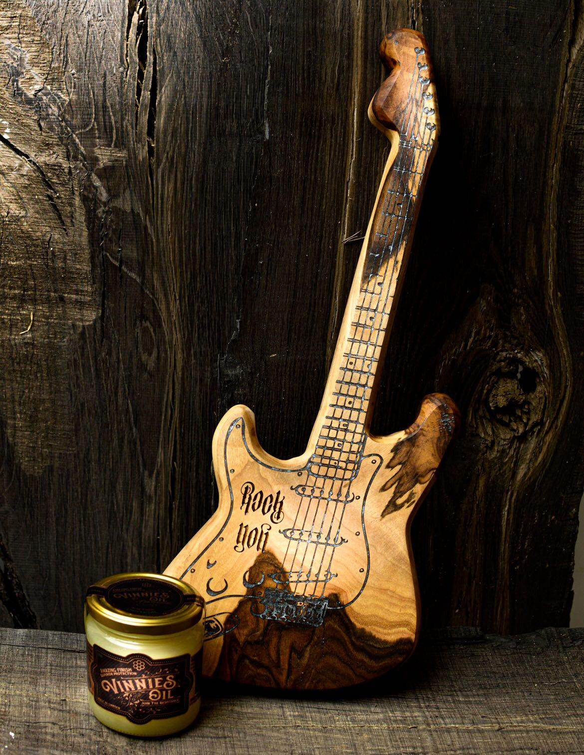 Wooden guitar cutting board with a jar of Vinnies Oil on a wooden surface.