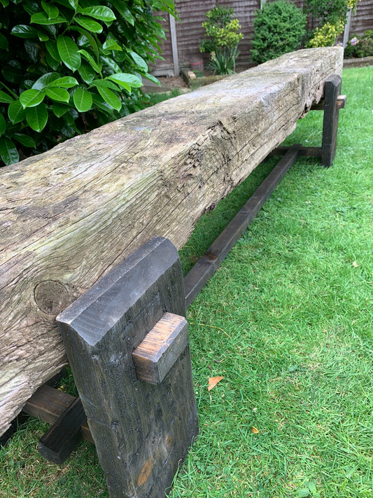 A dark oak finished garden sleeper bench leg placed on grass with a background of greenery and a traditional rustic aged sleeper.