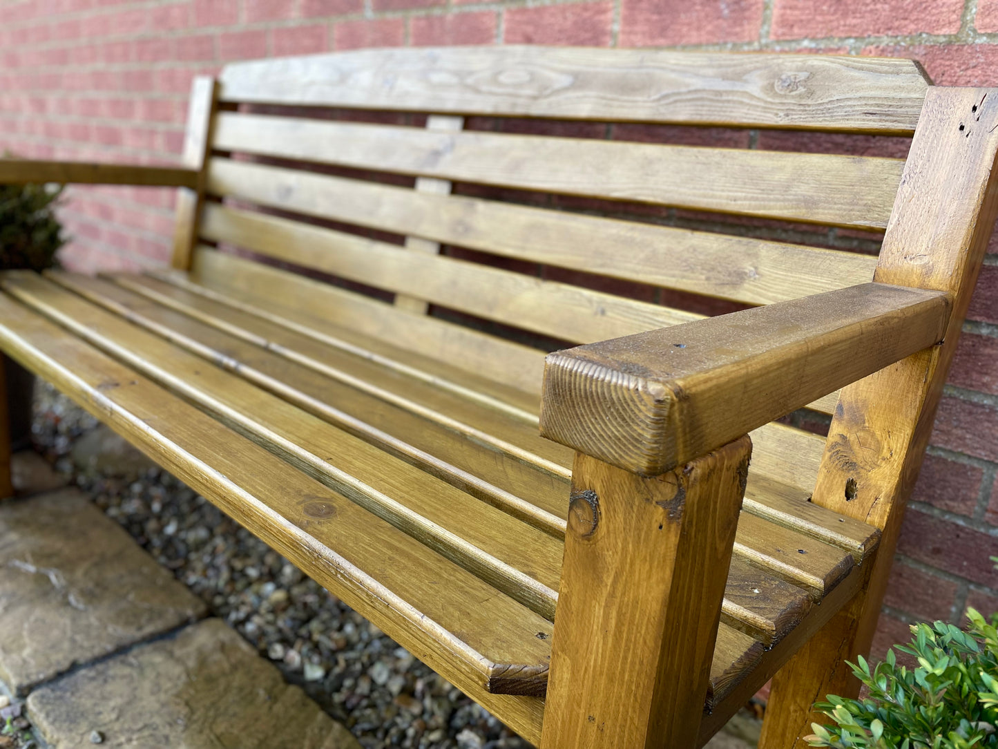 A medium oak stained wooden garden bench with visible wood grain, sitting against a brick wall with plants beside it.